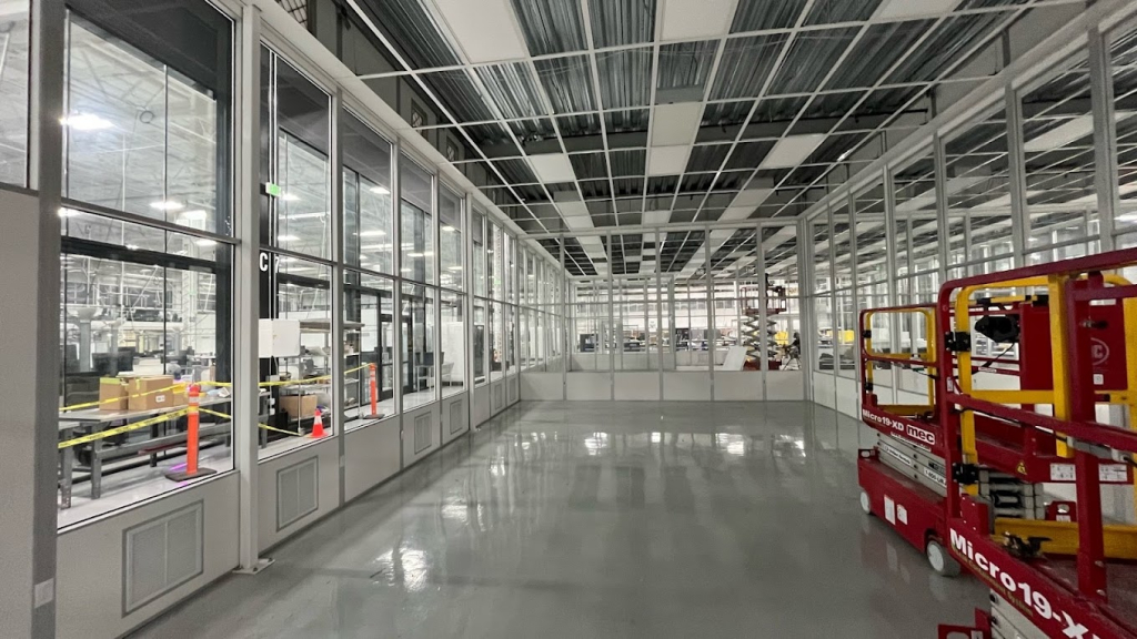 Modular cleanroom under construction inside a manufacturing facility, featuring glass walls, metal framing, and industrial equipment.