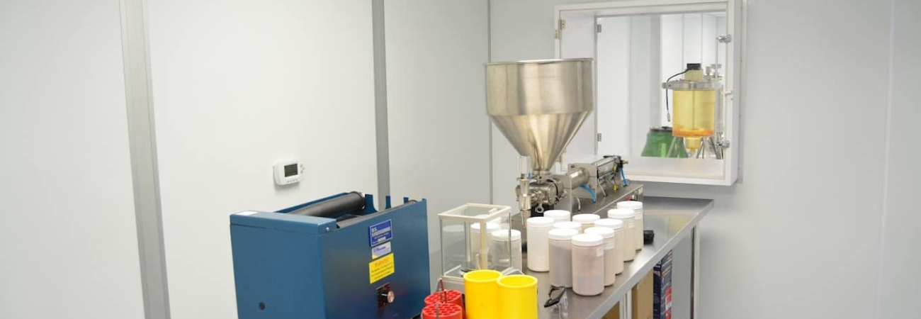 A cleanroom with sterile white walls, a blue machine, a large metal hopper, and various containers on a metal table, adhering to cGMP or GMP standards for pharmaceutical or chemical manufacturing.