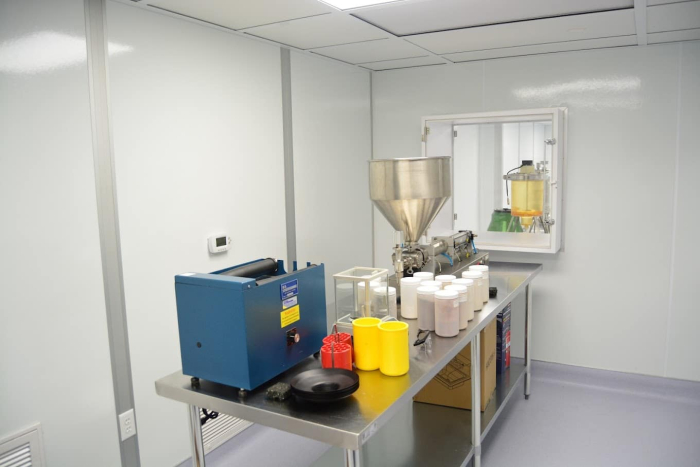 Cleanrooms A cleanroom with sterile white walls, a blue machine, a large metal hopper, and various containers on a metal table, adhering to cGMP or GMP standards for pharmaceutical or chemical manufacturing.